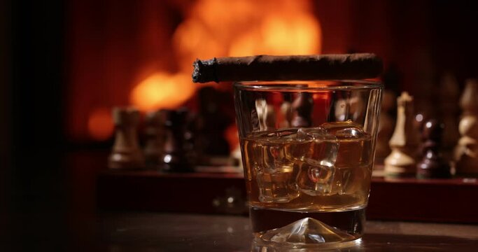 Whiskey and steaming cigar on the wooden weathered table in front of a fireplace. Glass of alcohol against the background of flames. Magical relaxed cozy atmosphere near fire.