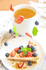 piece of strawberry and blueberry cheescake on plate and cup of tea served for breakfast at home, hotel or cafe. Berries levitating and falling down. Dessert meal, sweet food . High quality photo.