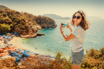 Fototapeta na wymiar Tourist girl taking photos on smartphone of the scenic beach Buyukcakil in resort town of Kas in Turkey. Sightseeing and travel blogger concept