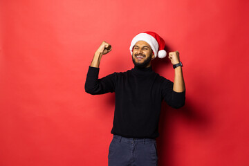 Young indian man in Santa hat with win gesture standing isolated on red background