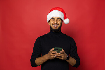 Young indian man in Santa hat use phone send greeting online standing isolated on red background