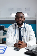 Portrait of african american practitioner doctor sitting at desk in hospital office. Therapist man working at patient sickness expertise analyzing medical documents. Heathcare service