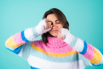 Young woman in a bright multicolored sweater on a blue background sleepy eyes, rubs her eyes with her hands, lack of sleep