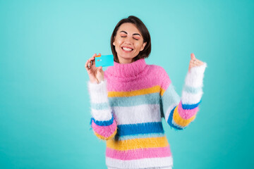 A young woman in a bright multicolored sweater on a blue background shows a credit card, jumps with...