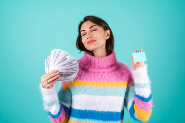 Young woman in a bright multicolored sweater on a blue background shows a credit card and a bundle...