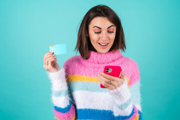 Young woman in a bright multicolored sweater on a blue background holds a credit card, pays for...