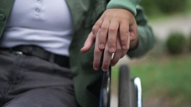 Unrecognizable boy holding hand of disabled girl on wheelchair armrest. Close-up palms of caring empathetic child and paralyzed friend outdoors in summer park. Support and friendship