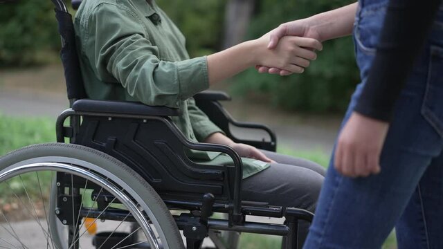 Side view unrecognizable disabled Caucasian girl greeting friend shaking hands in slow motion. Paraplegic ill teenager in wheelchair meeting friend outdoors in summer park