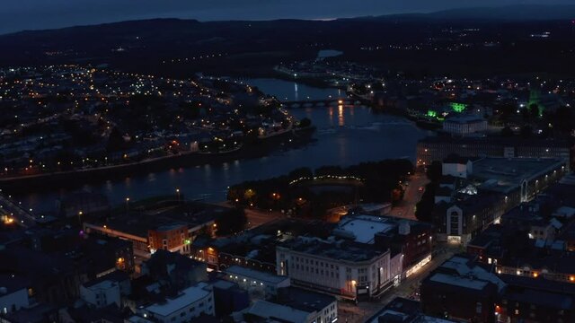 Fly above city in evening. Aerial view of river flowing through night town with street lights. Limerick, Ireland