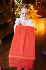 High-angle view of adorable smiling little blonde child girl sending gift box with Christmas present to you looking at camera in cozy living room, background of wall decorated xmas bokeh lights.