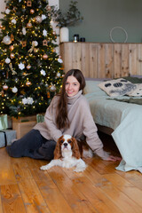 Happy young woman with King Charles spaniel dog at home with new year tree.