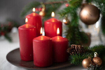 Christmas decoration with five red candles on brown plate, spruce branches, cones, balls and garland on white table on blurry background. Side view. New year mood, festive concept, holiday table.