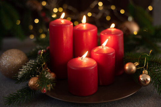 Christmas decoration with red candles on brown plate, spruce branches, cones and balls on gray table on background shining light. Side view. New year mood, festive concept, holiday table, gift card.