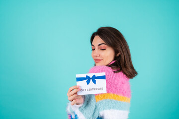 Young woman in a bright multicolored sweater on a blue background holds a gift certificate, smiles enthusiastically, beautiful makeup, nude plump lips