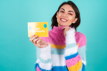 Young woman in a bright multicolored sweater on a blue background holds a gift certificate, smiles...
