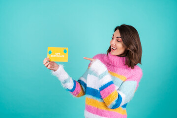 Young woman in a bright multicolored sweater on a blue background holds a gift certificate, smiles enthusiastically, beautiful makeup