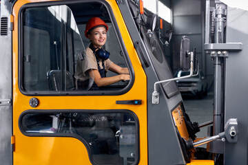 Fototapeta na wymiar Female worker driving loader in industrial container warehouse, looking happy, dressed in engineer uniform and hardhat orange helmet, smiling at camera. agriculture concept