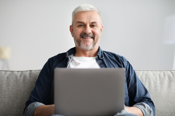 Fototapeta premium Cheerful mature man using laptop while resting on sofa