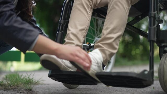 Unrecognizable woman adjusting legs of disabled child on wheelchair legrest in slow motion. Young Caucasian mother taking care of paraplegic son outdoors in summer park