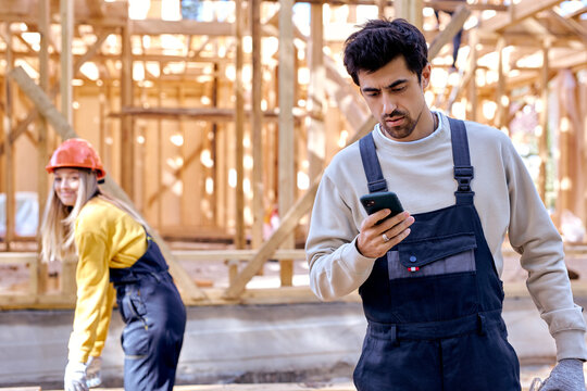 bearded caucasian male constructor in working uniform use smartphone reading message, in unfinished construction site, work place. successful cooperation of two builders, outdoors.