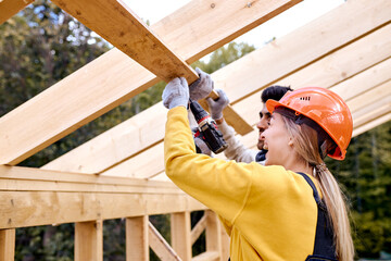 Two Young Contractors Working With Drill Driver. Carpenter Working on Wooden House Skeleton Frame Roof Section. Construction Industry Theme. Female In Hardhat Is Laughing During Work