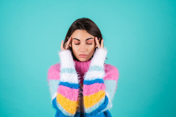 Young woman in a bright multi-colored sweater on a blue background, suffering from a headache,...