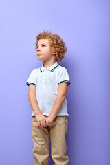 Shy preschool boy posing isolated over purple studio background. Curly kid in casual clothes looking up. Concept of facial expressions, childhood, education, family. Copy space for ad