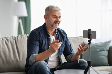 Attractive mature man broadcasting from home, using smartphone on tripod