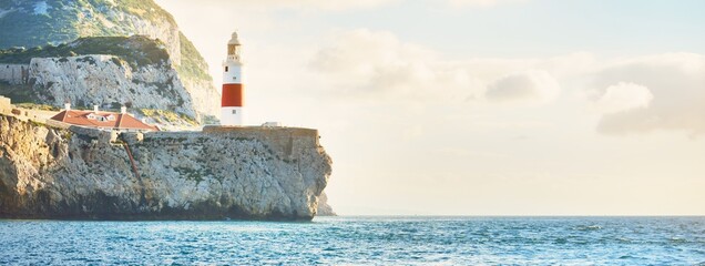 Trinity lighthouse at the rocky shore (cliffs) of the Europa Point. British Overseas Territory of Gibraltar, Mediterranean sea. Epic cloudscape. National landmark, sightseeing, travel destination - obrazy, fototapety, plakaty