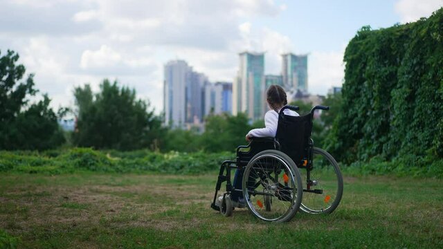 Wide shot summer park with disabled Caucasian girl in wheelchair admiring beauty of urban city at background. Back angle view paralyzed child alone outdoors looking away thinking. Slow motion