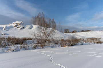 Winter landscape with a frozen river, covered with even clean snow, on which traces of sleds are visible. High rocky beach with a lonely wooden house. River valley with bushes and trees. Ural, Russia