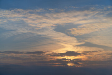 Sunset sky with white ornate clouds, tinted pink and yellow by the sun. A dark cloud has created a horizontal area, from under which the setting sun is breaking through. Tender patches of blue 