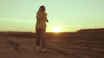 girl in headphones runs at sunset sky, listening to music for active training, speakers in ears of...