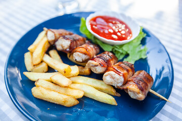 Chicken skewers with bacon. French fries and tomato sauce. Served on plate at restaurant table.