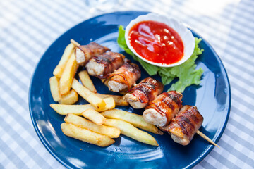 Chicken skewers with bacon. French fries and tomato sauce. Served on plate at restaurant table.