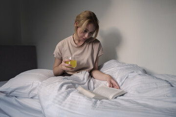 young blonde woman reading a book in bed and drinking orange juice, happy woman enjoying in bed on sunny morning