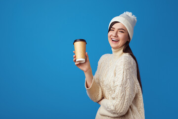Joyful caucasian female in white hat and white warm sweater, holds takeaway cup of coffee, poses against blue background