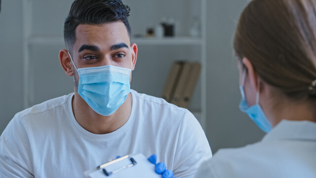 Arabic Man Hispanic Guy Patient Sick Male Wears Medical Protective Mask On Face Talks About Symptoms Of Health Problems To Unrecognizable Doctor Woman Nurse Writing Down Notes, Consultation In Clinic.