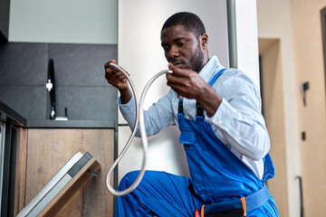 Young African Handyman Repairing Dishwasher, Need To Change Old Dishwasher Hose, Black Guy In Blue...