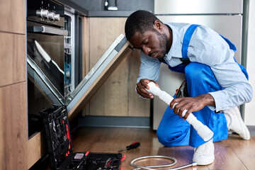 Side View Young African Handyman Repairing Dishwasher, Changing Siphon, Wearing Blue Workwear Overalls. Confident Professional Handyman Ar Home ,Focused On Work Alone, In Kitchen
