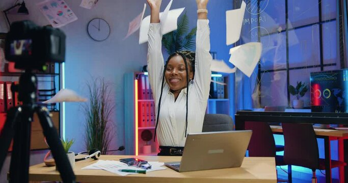 Beautiful satisfied successful african american woman with stylish hairstyle holding online chat with her internet audience and throwing up sheets of papers in the end of stream