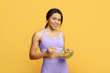 Healthy food concept. Fit african american woman eating fresh vegetable salad, smiling to camera over yellow background