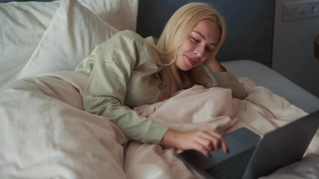 Cheerful blond woman looking at laptop in bed at home
