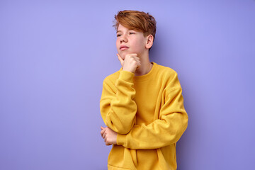 Let me think. Portrait of pensive clever redhead schooler in yellow shirt with puzzled serious expression, child thinking doubting, making choice. indoor studio shot isolated on purple background