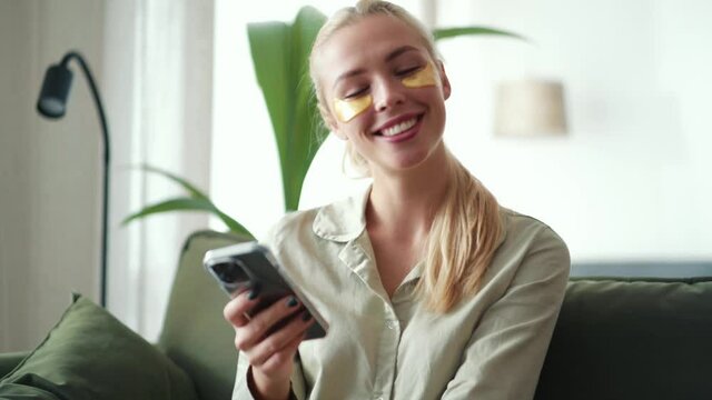Serious blond woman with sticking patches to eyes texting by phone at home