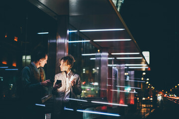 Office worker man shows woman colleague information on smartphone and discusses project standing on transport stop against city traffic in late evening