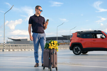 A young businessman in a parking lot with luggage and flowers calls a taxi service by phone. Business trip