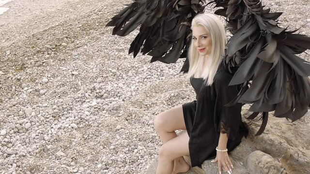 Sexy Blond Young Woman Dressed As Angel With Black Wings Sitting On The Stone On The Beach. Fashion Female Model Smiles Looking At Camera . High Angle View.