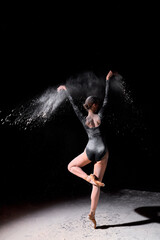 gorgeous slender ballet dancer woman wearing bodysuit, posing dancing among the cloud of flying flour on black studio background, make performance. Artistic, commercial, monochrome design.