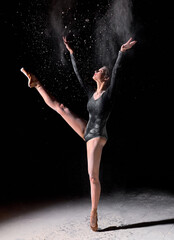 Fototapeta graceful slim woman in black bodysuit emotionally dancing on black background in clouds of white dust, in studio. ballerina female in slow motion action, indoors, performance by young lady obraz